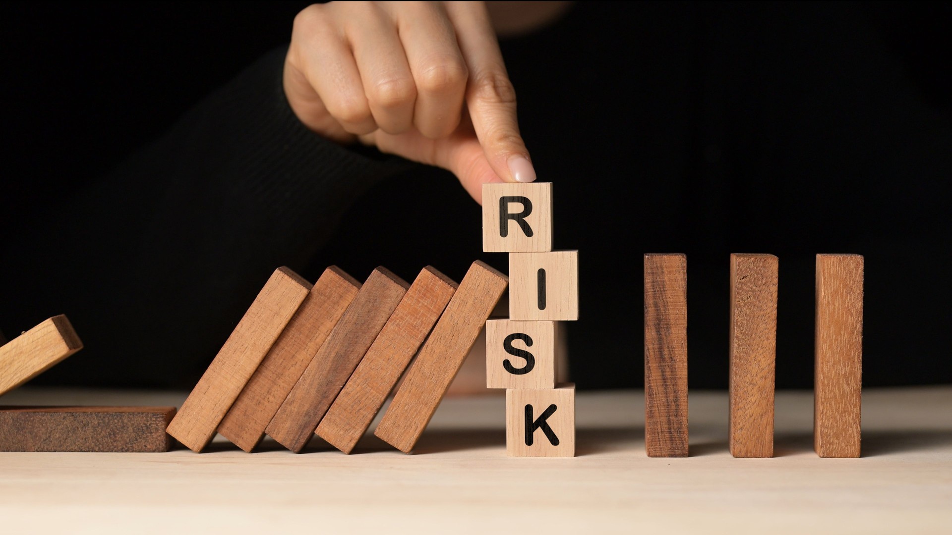 A close-up of a woman's hand balancing wooden blocks labeled "RISK," falling dominos nearby, symbolize risk management and mitigation in business through strategic actions